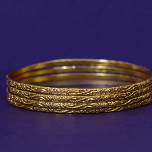 May include: Four gold-coloured bangles with intricate, embossed designs. The bangles are stacked on top of each other.