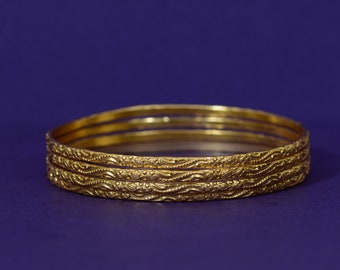 May include: Four gold-coloured bangles with intricate, embossed designs. The bangles are stacked on top of each other.