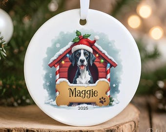 May include: A white, round Christmas ornament with a dog wearing a Santa hat inside a red doghouse. The dog's name, "Maggie", is on a bone-shaped tag. The ornament has a white ribbon for hanging and the year 2025 is printed on the bottom.