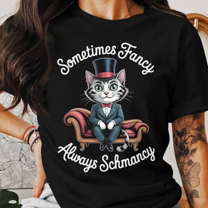 Op de afbeelding: Zwart t-shirt met een cartoonkat in smoking en hoge hoed, zittend op een rode bank. De tekst "Sometimes Fancy, Always Schmancy" staat boven en onder de kattenillustratie. De kat heeft groene ogen en een vlinderdas.