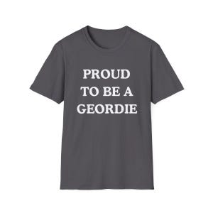 May include: A dark gray short-sleeved t-shirt featuring the white text PROUD TO BE A GEORDIE printed in a bold, sans-serif font across the center of the chest. The garment has a classic crew neckline and a straight hemline, providing a simple design.