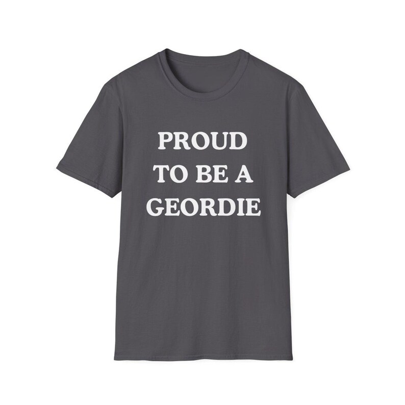 May include: A dark gray short-sleeved t-shirt featuring the white text PROUD TO BE A GEORDIE printed in a bold, sans-serif font across the center of the chest. The garment has a classic crew neckline and a straight hemline, providing a simple design.