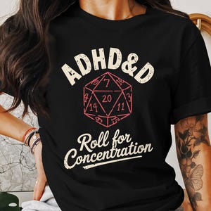 Könnte beinhalten: Schwarzes T-Shirt mit weißem Text, der "ADHD&D" lautet, und einer Grafik eines roten 20-seitigen Würfels. Unter dem Würfel steht der Text "Roll for Concentration".