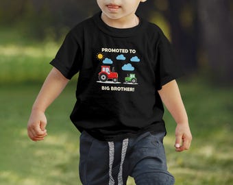 May include: Black t-shirt with a graphic featuring a red tractor, a green tractor, a sun, and clouds. Text reads "Promoted to Big Brother!" A fun design for a new big brother.