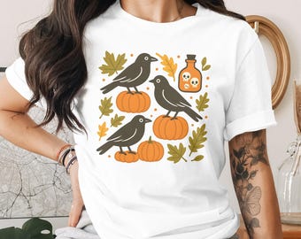 Peut inclure: T-shirt blanc avec un motif sur le thème d'Halloween. Le graphisme comprend trois corbeaux noirs perchés sur des citrouilles oranges, entourés de feuilles d'automne et d'une bouteille de potion avec des icônes de crâne. Un design festif.