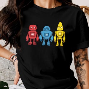 以下が含まれることがあります： 黒いTシャツに、3体のカラフルなロボットのイラストが描かれています。ロボットは赤、青、黄色で、それぞれ異なるデザインと番号（01、10、11）がついています。デザインはTシャツの中央に配置されています。