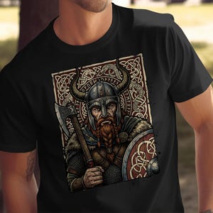 May include: A black t-shirt featuring a graphic print of a bearded warrior wearing a horned helmet, holding an axe and a round shield. The design includes intricate knotwork patterns and runic inscriptions set within a rectangular frame.