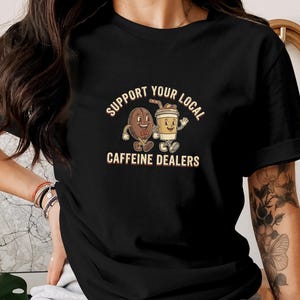 以下が含まれることがあります： 「SUPPORT YOUR LOCAL CAFFEINE DEALERS」の文字と、コーヒー豆とコーヒーカップの漫画のグラフィックがプリントされた黒いTシャツ。 ヴィンテージスタイルです。