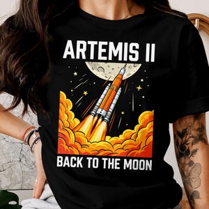 May include: Black t-shirt featuring a graphic of a rocket launching towards the moon, with the text "ARTEMIS II" and "BACK TO THE MOON" in white. The design includes orange flames and a starry sky.