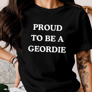 May include: A black short-sleeved t-shirt featuring the white text PROUD TO BE A GEORDIE printed in a bold, sans-serif font across the chest. The garment has a classic crew neckline and a standard fit, showcasing the text clearly against the dark fabric.