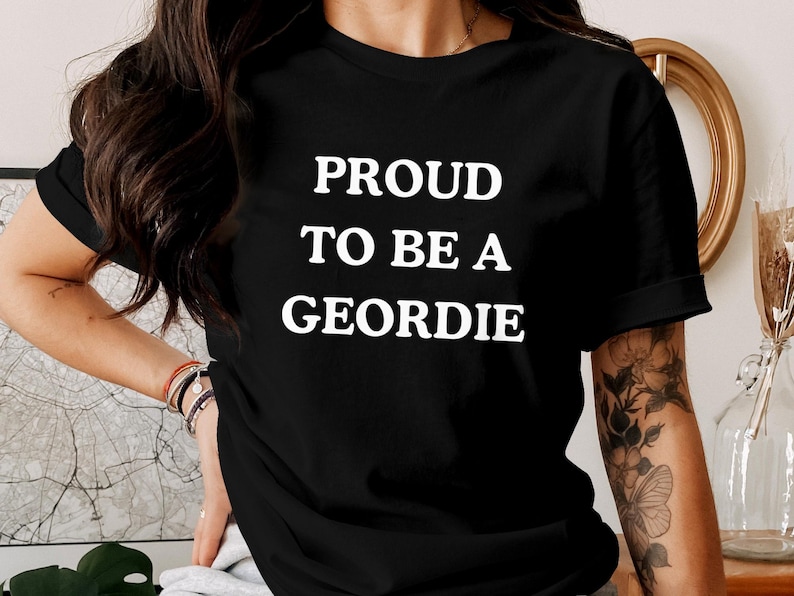 May include: A black short-sleeved t-shirt featuring the white text PROUD TO BE A GEORDIE printed in a bold, sans-serif font across the chest. The garment has a classic crew neckline and a standard fit, showcasing the text clearly against the dark fabric.