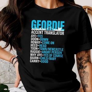 Puede incluir: Camiseta negra con el texto "GEORDIE ACCENT TRANSLATOR" en azul y blanco. La camiseta enumera t&eacute;rminos de jerga Geordie y sus traducciones al ingl&eacute;s. Una camiseta gr&aacute;fica divertida.