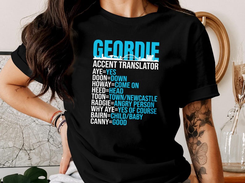 Puede incluir: Camiseta negra con el texto "GEORDIE ACCENT TRANSLATOR" en azul y blanco. La camiseta enumera t&eacute;rminos de jerga Geordie y sus traducciones al ingl&eacute;s. Una camiseta gr&aacute;fica divertida.