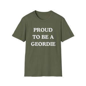 May include: An olive green short-sleeved t-shirt featuring the white text PROUD TO BE A GEORDIE printed in a bold, sans-serif font across the center of the chest. The garment has a classic crew neckline and a straight hemline at the bottom.