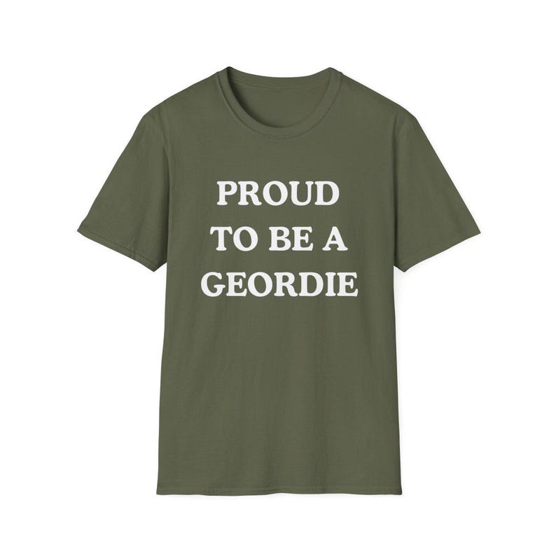 May include: An olive green short-sleeved t-shirt featuring the white text PROUD TO BE A GEORDIE printed in a bold, sans-serif font across the center of the chest. The garment has a classic crew neckline and a straight hemline at the bottom.