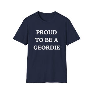 May include: A navy blue short-sleeved t-shirt featuring the white text PROUD TO BE A GEORDIE printed in a bold, sans-serif font across the center of the chest. The garment has a classic crew neckline and a straight hemline design.