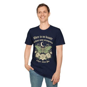 May include: Navy blue t-shirt featuring a green moth design with a crescent moon and floral accents. The shirt includes the quote "There is no beauty without some strangeness" by Edgar Allan Poe.