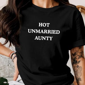 Puede incluir: Camiseta negra con el texto blanco "HOT UNMARRIED AUNTY". Esta prenda informal es perfecta para el uso diario. La imagen muestra la camiseta en una persona.