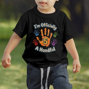May include: Black t-shirt with the text "I'm Officially A Handful" and a large orange handprint with the number 5. Surrounding the handprint are smaller handprints in blue, red, green, and purple. The shirt is worn by a child.