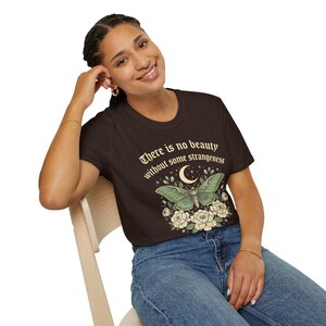 May include: A brown t-shirt featuring a green moth design with a crescent moon and floral accents. The shirt has the text "There is no beauty without some strangeness" in a decorative font. The model is wearing blue jeans.