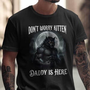 May include: Black t-shirt with a werewolf illustration under a full moon. The text "DON'T WORRY KITTEN" is above the image, and "DADDY IS HERE" is below. The design is printed on a black t-shirt.