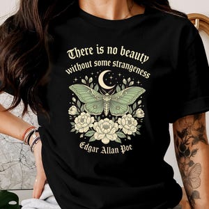 May include: Black t-shirt featuring a green moth design with a crescent moon and floral accents. The text "There is no beauty without some strangeness" and "Edgar Allan Poe" are also displayed.