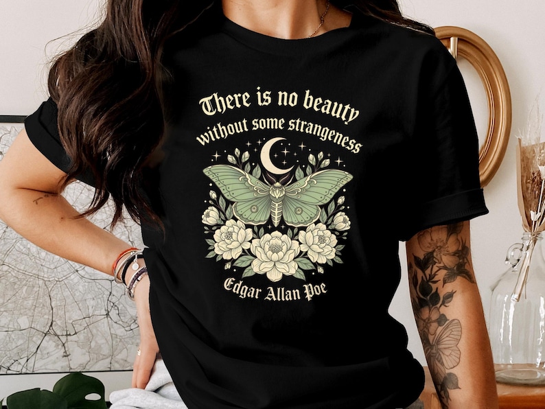 May include: Black t-shirt featuring a green moth design with a crescent moon and floral accents. The text "There is no beauty without some strangeness" and "Edgar Allan Poe" are also displayed.