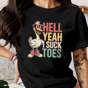 May include: Black t-shirt with a cartoon goose wearing sunglasses and red trainers. The shirt features the text "HELL YEAH I SUCK TOES" in a retro font. The goose graphic is the main design element.