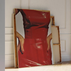 May include: A framed print of a woman wearing a red leather dress. The print is leaning against a white wall.
