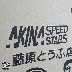 May include: Dark grey sticker featuring the text "AKINA SPEED STARS" in a bold font. Japanese characters are below. The sticker is set against a white background, with a small "Sticky Man" logo.