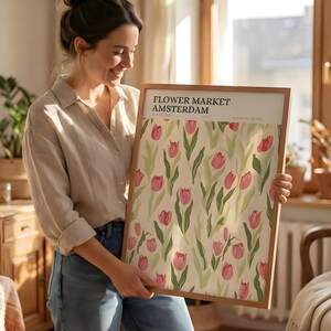 Woman smiling and holding a large framed pastel flower market Amsterdam poster in a bright, sunlit room.