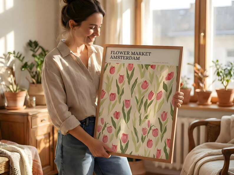 Woman smiling and holding a large framed pastel flower market Amsterdam poster in a bright, sunlit room.