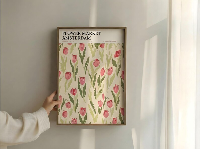 Hand adjusting a black-framed Amsterdam flower market exhibition poster hanging on a beige wall with soft natural light.