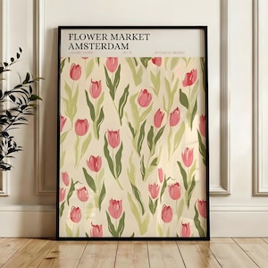Framed Amsterdam flower market poster featuring pink tulips, leaning against a white wainscoted wall next to a plant.
