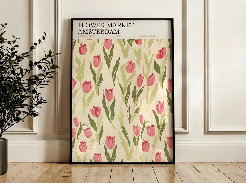 Framed Amsterdam flower market poster featuring pink tulips, leaning against a white wainscoted wall next to a plant.