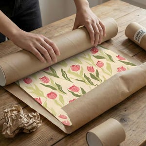 Hands carefully unrolling a large vintage-inspired Amsterdam flower market poster with pink tulips on a wooden table.