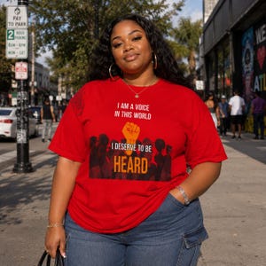 Op de afbeelding: Een felrood T-shirt met korte mouwen en een grafische print van een opgeheven vuist boven een menigte mensen. De tekst I AM A VOICE IN THIS WORLD en I DESERVE TO BE HEARD staat in witte en oranje letters op de voorkant van de borst.