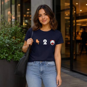 May include: A navy blue short-sleeved t-shirt featuring a graphic print on the chest. The design consists of an orange finger pointing down, the white number two, and a colorful clown face emoji arranged in a horizontal line across the front.