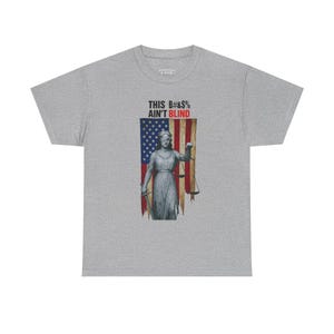 May include: A heather gray t-shirt featuring a graphic of Lady Justice with the American flag as a backdrop. The text "THIS B#&%S% AIN'T BLIND" is printed above the image. The shirt is made of a soft, comfortable material.