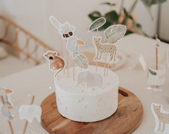 May include: A white cake decorated with jungle animal and leaf themed cake toppers. The cake is on a wooden cake stand.