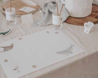 May include: A white placemat with a blue whale, a brown stingray, a seagull, and brown seashells printed on it. The placemat is on a beige tablecloth.