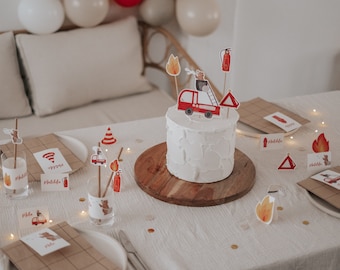 May include: A white cake decorated with a red fire truck, fire hydrant, and flames. The cake is on a wooden cake stand and surrounded by red and yellow fire-themed decorations. The table is covered in a white tablecloth with gold confetti.