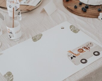 May include: A white table setting with a white cake, a wooden tray with blueberries, and a place setting with a white placemat featuring a jungle animal bus design. The placemat has a green leaf design and the words "Matilda" written on it.