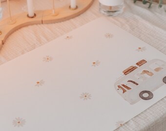 May include: A white placemat with a light brown daisy pattern and a watercolor illustration of a white bus with luggage on top. The bus is driving on a light brown road.