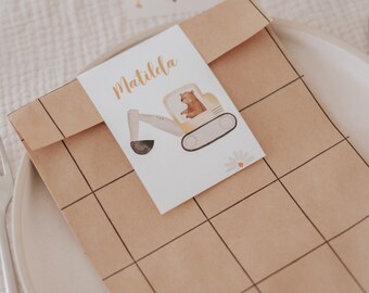 May include: A brown paper bag with a grid pattern, a white card with the name Matilda and a cartoon excavator with a bear inside, and a white card with the name Mila and a cartoon cement mixer.