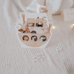 May include: A white card with a brown wooden ark illustration. The ark is filled with various animals, including a giraffe, a zebra, a lion, a monkey, and a bird. The card is on a white tablecloth with a few small white leaves scattered around it.