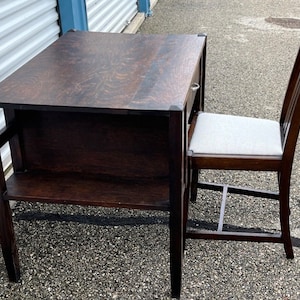May include: A dark brown wooden desk and chair set. The desk has a rectangular top, a lower shelf, and a small drawer. The chair has a light gray cushioned seat and a curved back. The set is made of wood and has a classic design.