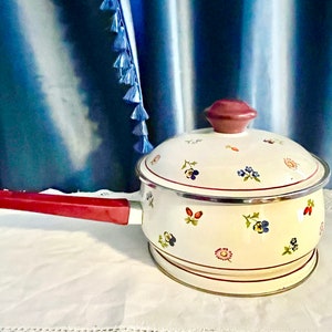 May include: Cream-colored saucepan with a lid, featuring a floral pattern of small flowers in red, blue, and yellow. The handle and lid knob are a deep red. The pan has a silver rim and sits on a white surface.