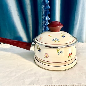 May include: A white enamel saucepan with a red handle and a floral design. The saucepan has a lid with a red knob and is sitting on a white surface.