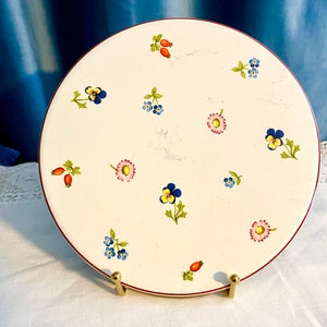 May include: A white ceramic plate with a red rim and a floral design. The plate features small blue, yellow, and pink flowers, as well as red berries.